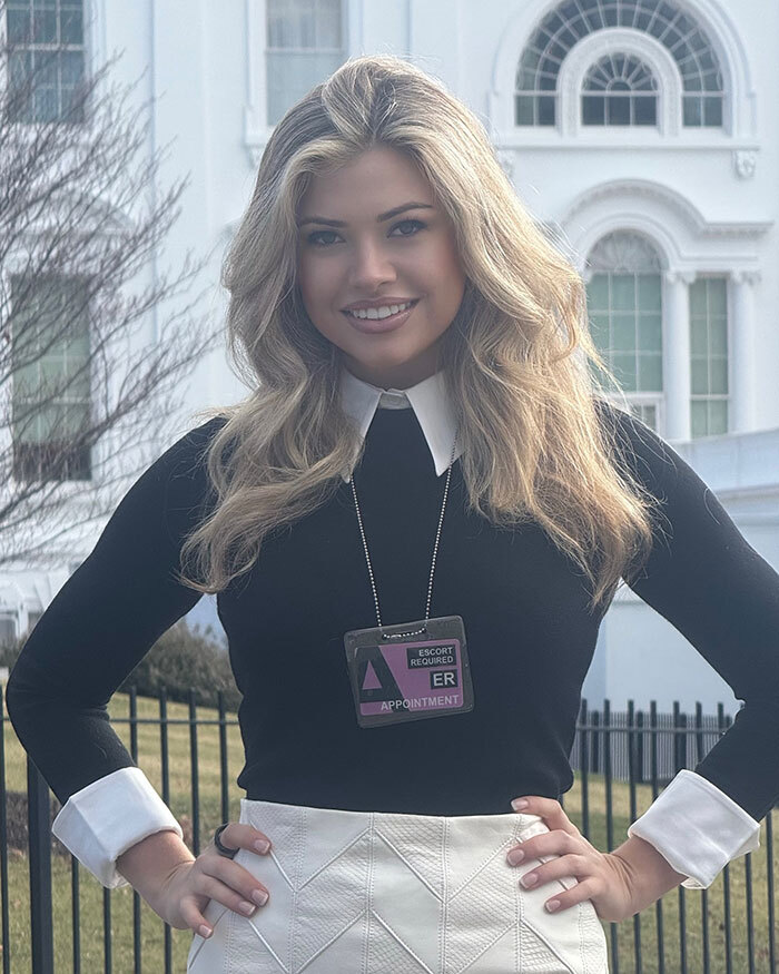 White House correspondent in a collared outfit outside the White House, wearing a badge around her neck.