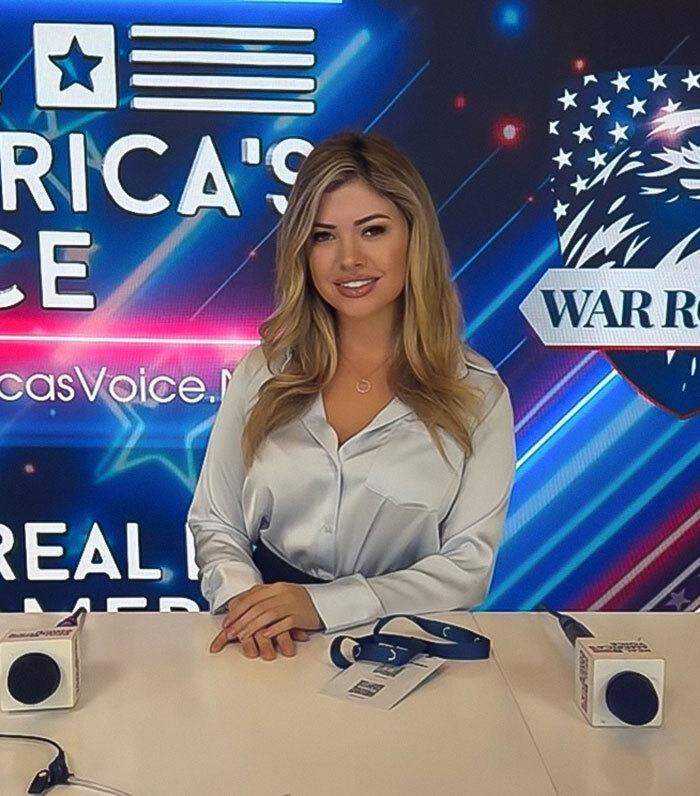 White House correspondent in a studio setting, wearing a light-colored blouse, with broadcasting equipment on the table.