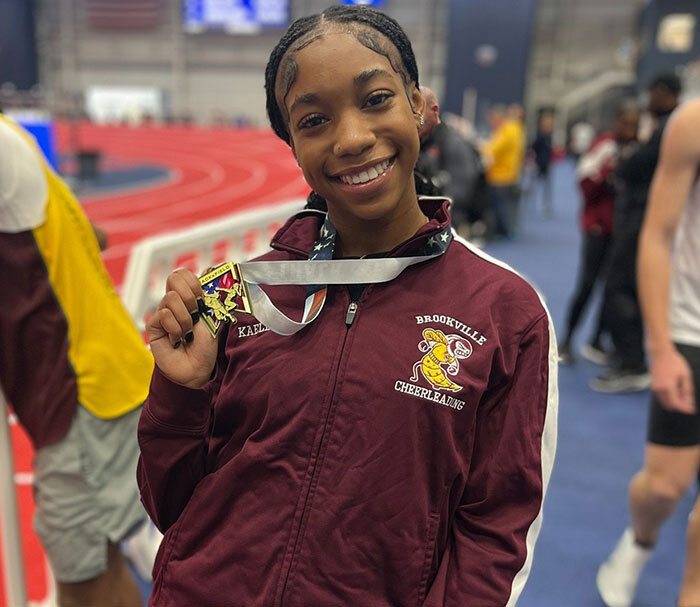 Runner smiling with a medal on an indoor track, wearing a maroon jacket with "Brookville Cheerleading" logo.