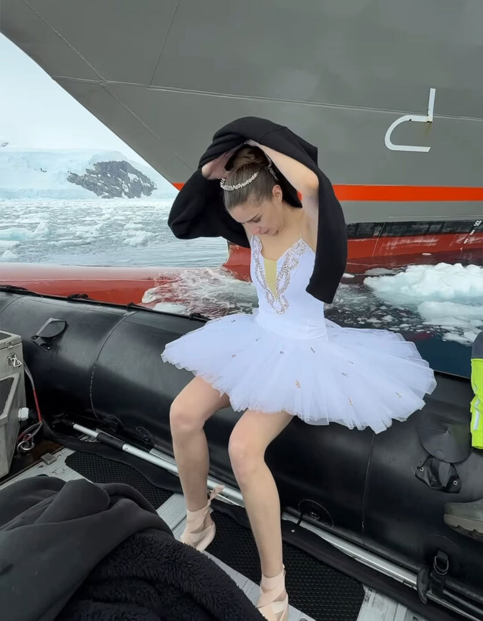 Ballerina wearing a tiara and tutu on a ship's bow in Antarctica waters, preparing to cover with a black jacket.