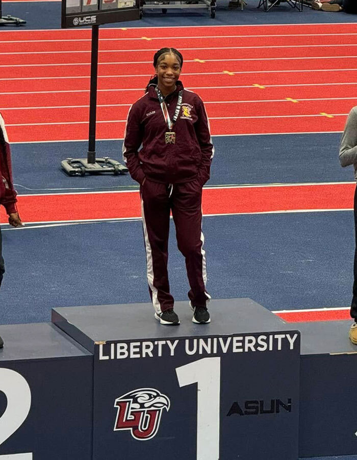 Athlete on podium at Liberty University track event, smiling and wearing a medal.