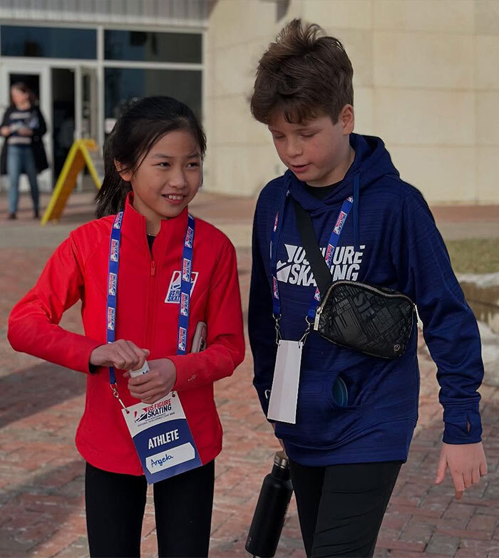 Child ice skating duo walking together, wearing athletic gear with badges.