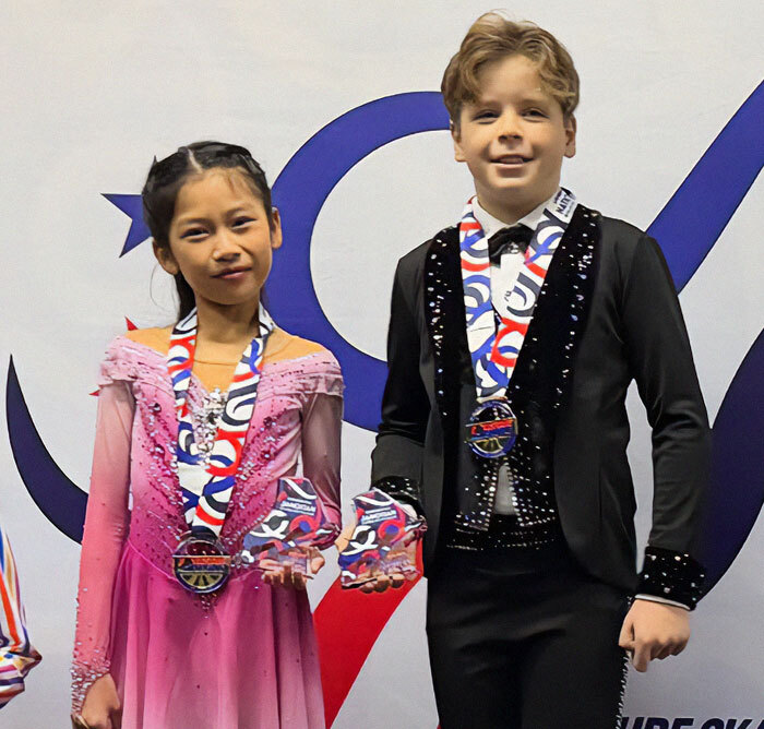 Child ice skating duo in costumes with medals, smiling after a performance.