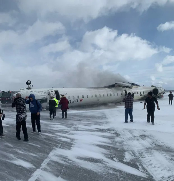 Delta Airlines plane upside down on snowy runway after crash landing, with people nearby.