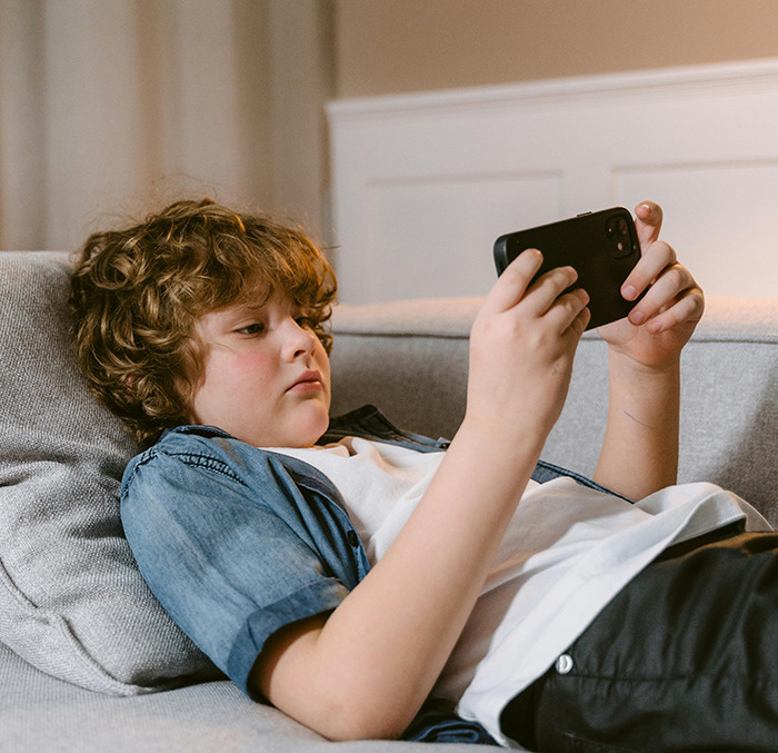 Child using a smartphone, reclining on a sofa in a casual setting.