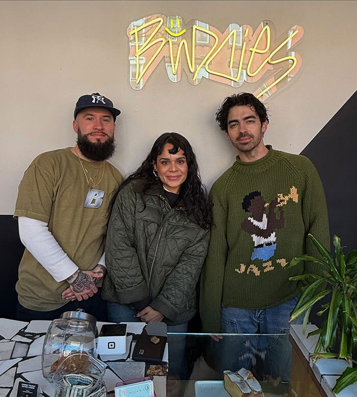 Three people in a NYC cafe, standing under a neon "Birdies" sign, with Joe Jonas among them.
