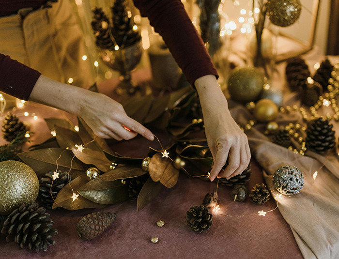 Hands arranging holiday decor with pine cones and lights on a festive table, creating a warm Christmas atmosphere.