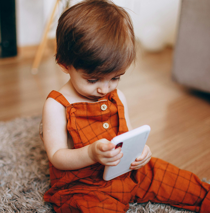 Toddler in orange overalls, engrossed in a smartphone while sitting on a carpet.