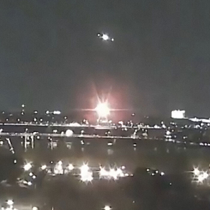 Nighttime scene with a helicopter flying over a city, lights illuminating the skyline amidst a Black Hawk drill.
