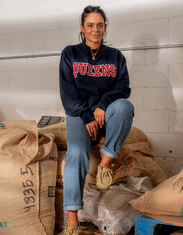 Smiling cafe owner sitting on coffee bags in NYC, wearing a "Queens" sweatshirt and jeans.
