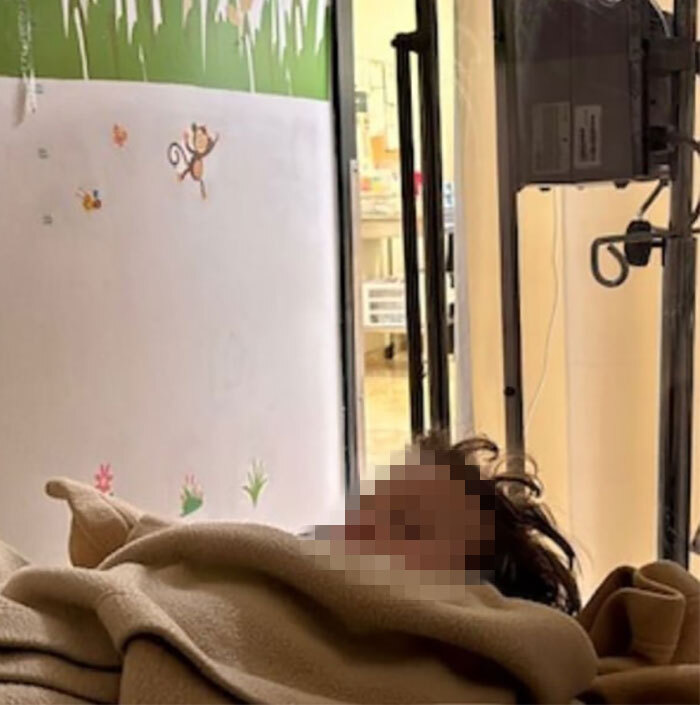 Person lying in a hospital bed, wrapped in a blanket, with a colorful mural on the wall.
