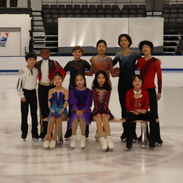 Teen skaters in colorful outfits pose on ice rink, some seated and others standing, before solemn event.