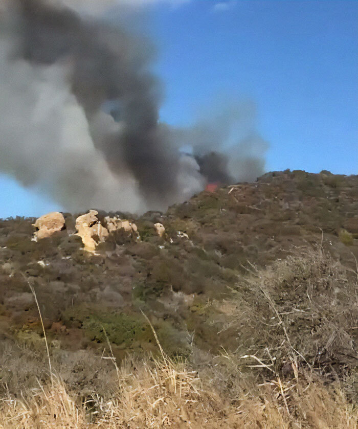 Thick smoke rises from a hillside fire in La Palisades under a clear blue sky.