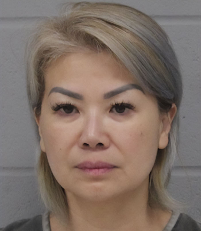 A woman under arrest related to mortuary misconduct, with a neutral expression, in front of a grey background.