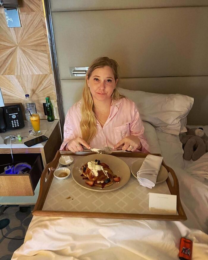 Person in a pink pajama top having breakfast in bed, with a tray of pancakes and drinks beside them.
