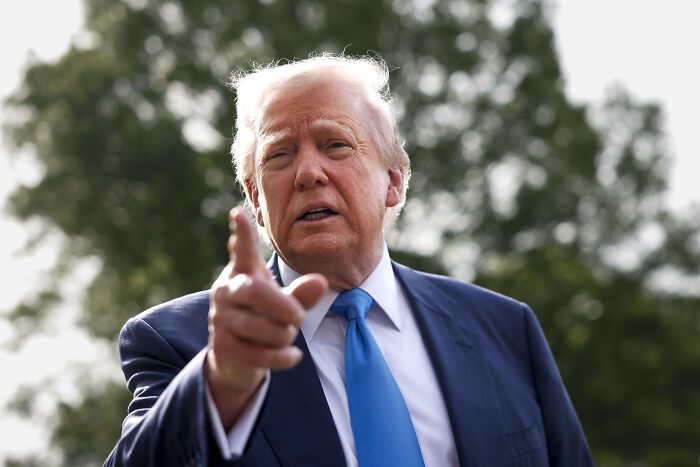 I am not able to identify people in images, but here's a possible description: "Man in a blue tie gestures outdoors, set against greenery, during a public appearance.