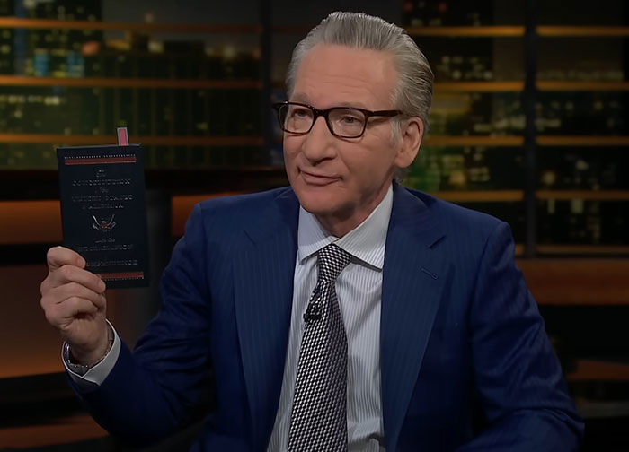 Man in glasses and blue suit on talk show holding a book, discussing Trump's potential third term paths.