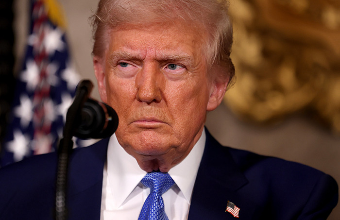 A person in a suit stands behind a microphone, appearing serious.