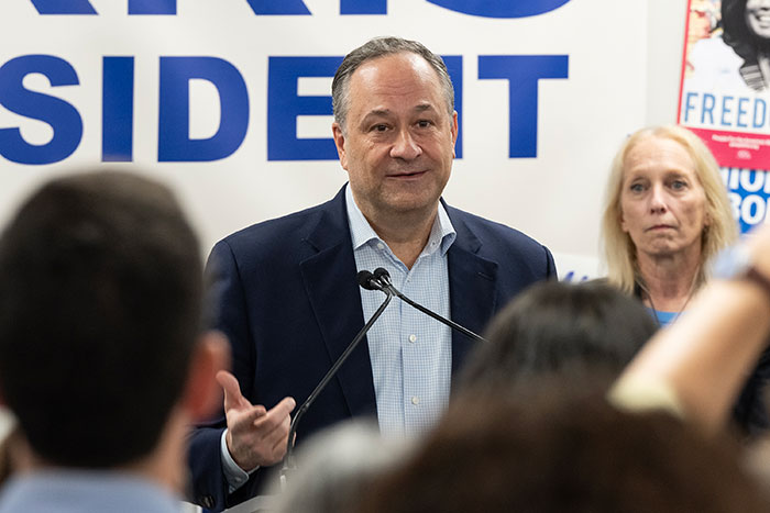 Man speaking at a podium during an event, with audience members in foreground and campaign signs in the background.