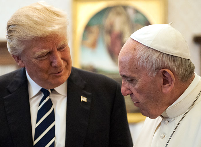 Trump in a dark suit speaking with a man dressed as pope, wearing a white papal skullcap and robes in a formal setting