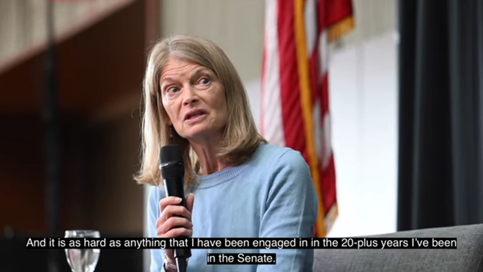 Senator discussing Trump&rsquo;s retaliation threat, holding a microphone, seated with an American flag in the background.