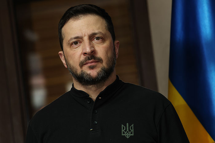 Man in a dark shirt with Ukrainian emblem standing in front of a flag, relating to Trump confusion and Russia Putin meeting.