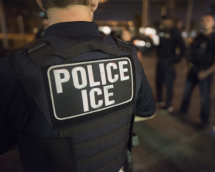 ICE police officer wearing a tactical vest with Police ICE patch standing with a group at night in a blurred background
