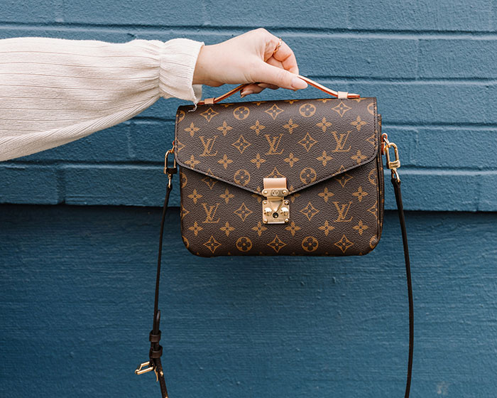 Hand holding a designer handbag in front of a blue wall, illustrating direct purchase appeal from factories to bypass tariffs.