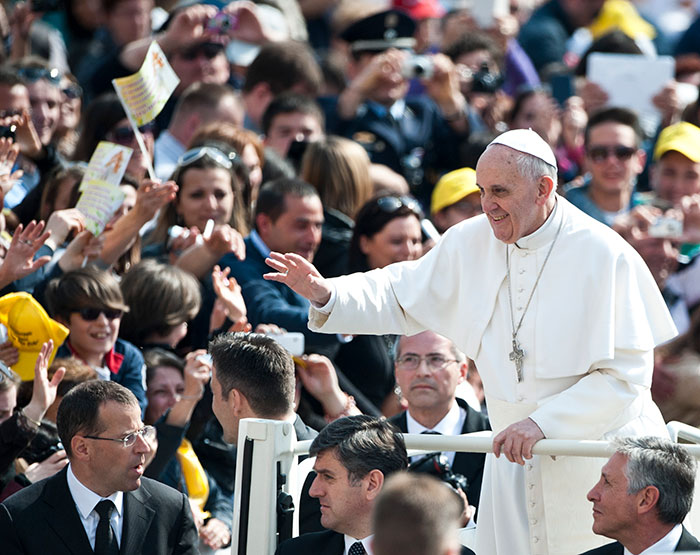 Pope greeting a crowd during a public event with people taking photos and waving in the background.