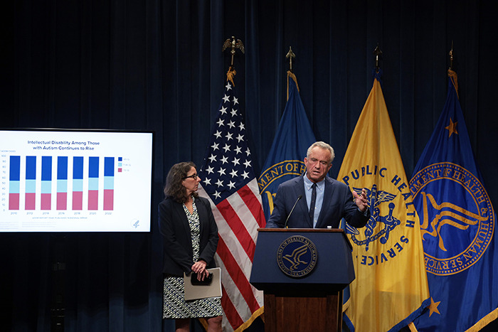 Man at a podium with flags, discussing autism registry plan, alongside a woman and a presentation slide with charts.