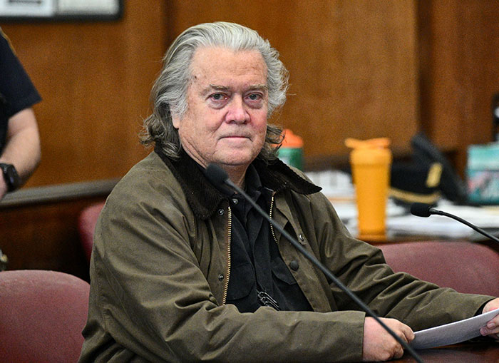 Ex-White House strategist sitting in a courtroom, holding papers, with a serious expression.