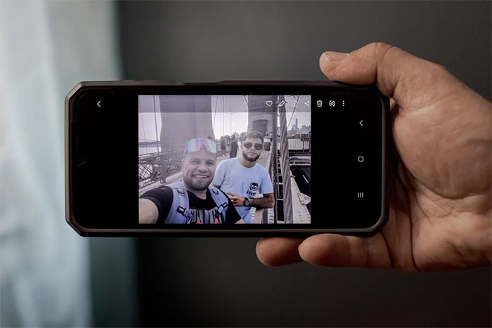 Hand holding a phone displaying a photo of two people smiling on a bridge, related to an ICE agents incident.