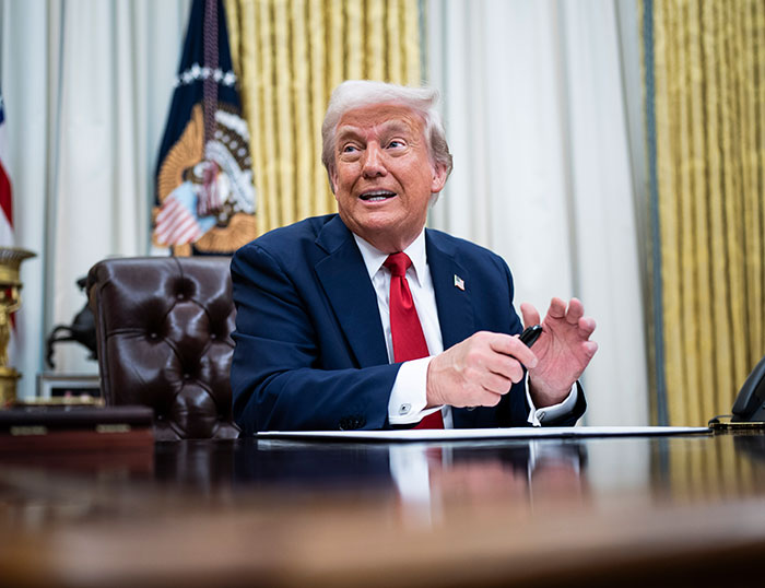 A man in a suit and red tie seated at a desk in a formal setting.