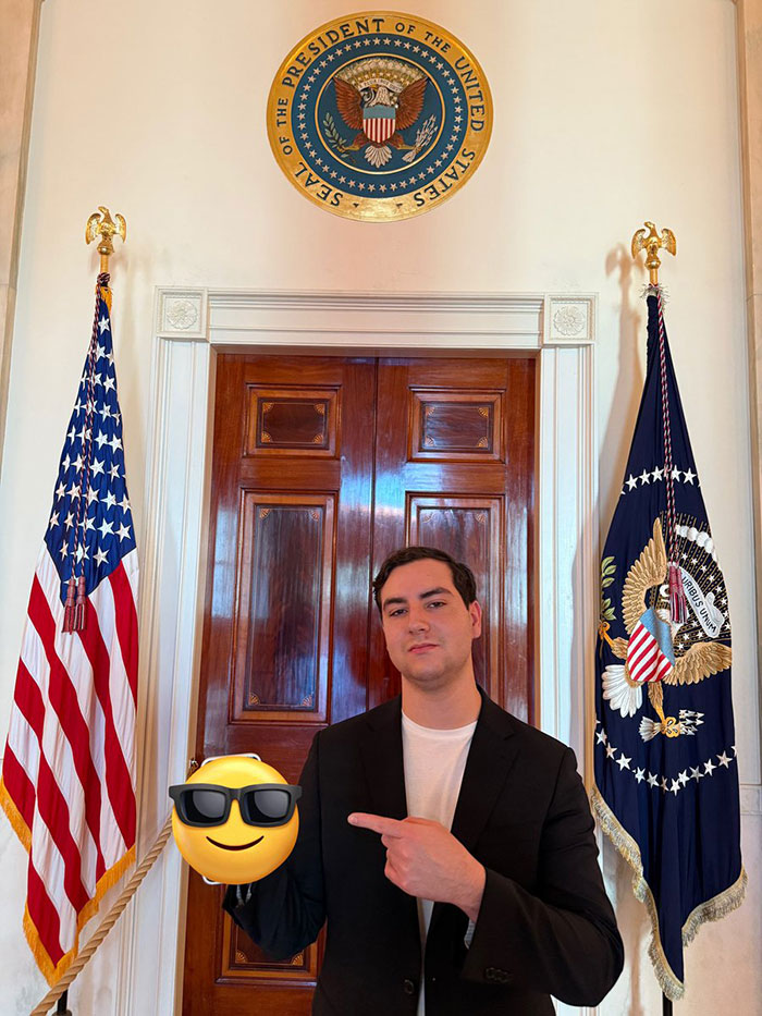 Man in black blazer posing in front of presidential seal with American flags, related to crypto bros Trump event.