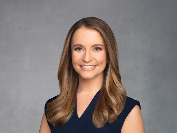 Young female reporter with long brown hair and a navy top, smiling in a professional studio setting discussing Trump TACO nickname.
