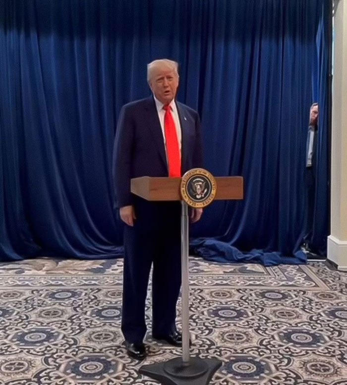 Former President Trump speaking at a podium with presidential seal in front of blue curtains during event.