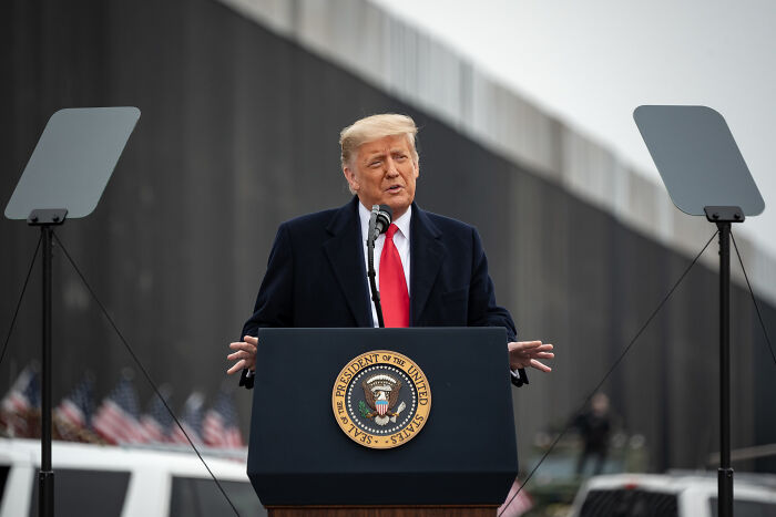 Donald Trump speaking at a podium with presidential seal, addressing audience near a border wall during an outdoor event.