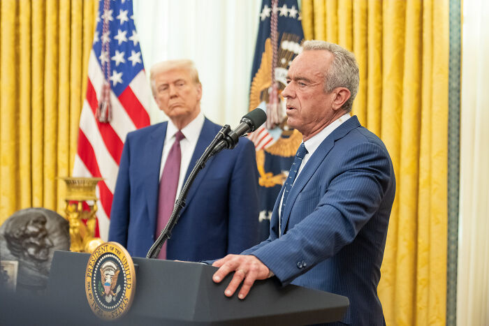 Health Secretary RFK Jr. speaking at a podium with the U.S. flag and President Trump in the background in the Oval Office.