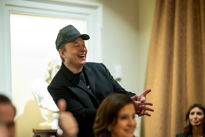 Elon Musk laughing and wearing a Make America Great Again hat during a casual indoor meeting with people.