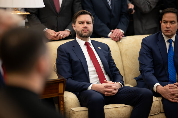 JD Vance seated in a suit with red tie, showing serious expression during event with Pope Leo in Rome.