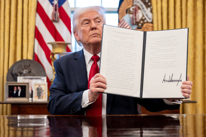 Former President Trump in the Oval Office holding a signed document, reacting to a reporter mentioning his TACO nickname.