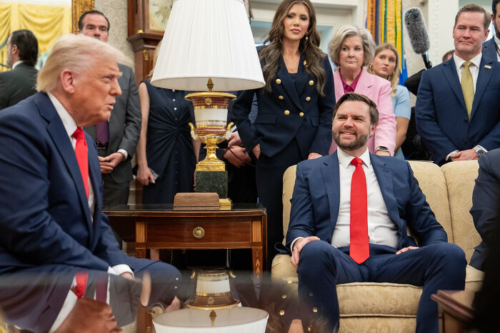 Donald Trump posing with red lightsaber on Star Wars Day, surrounded by people in a formal office setting.