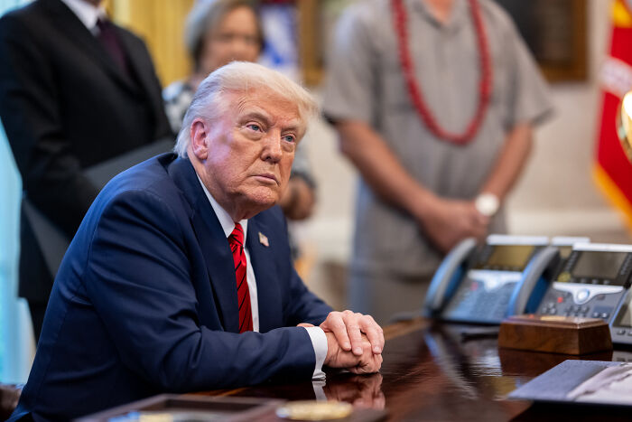 Donald Trump sitting at a desk in a suit and red tie, a key figure linked to Star Wars Day and red lightsaber discussions.