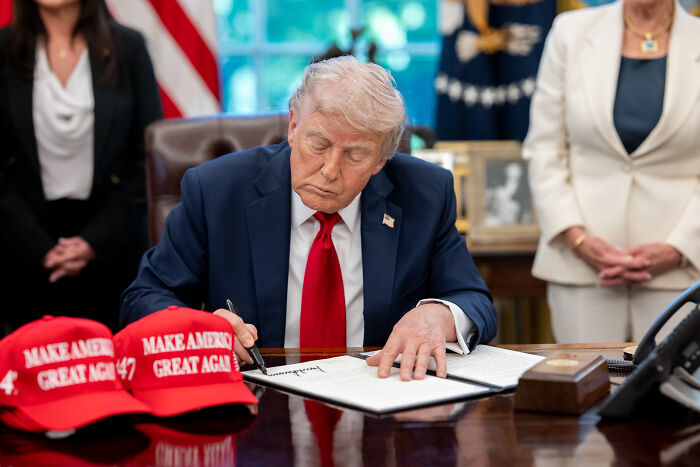 Donald Trump sitting at a desk signing documents, with red Make America Great Again hats and people standing nearby.