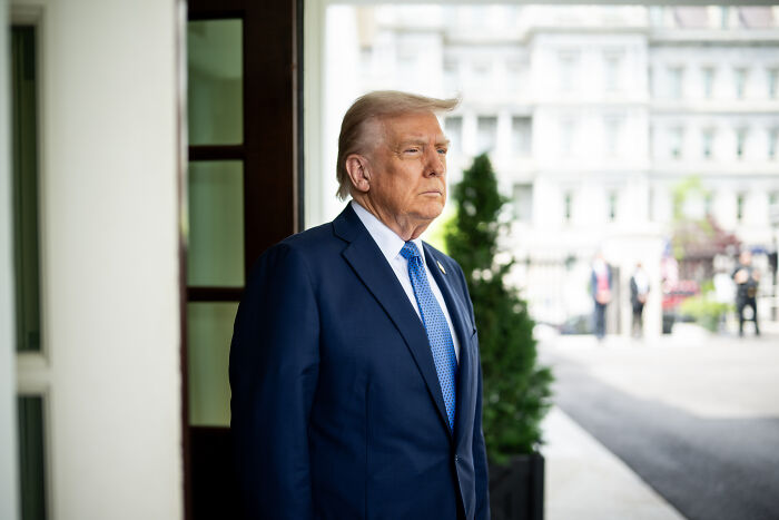 Donald Trump standing outside a building wearing a dark suit and blue tie, with a serious expression in daylight.