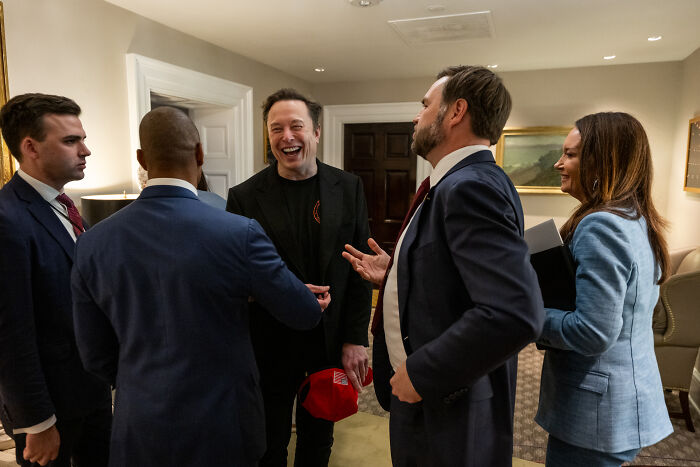 Elon Musk laughing and talking with a group of people in a formal room, discussing government waste and DOGE.