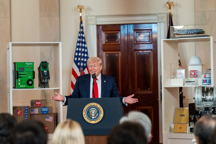 Donald Trump speaking confidently at a podium with presidential seal during a town hall event with an audience.