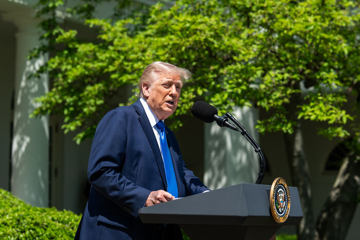 Former president Trump speaking at an outdoor podium with presidential seal, related to surgeon general nominee and wellness influencer news.