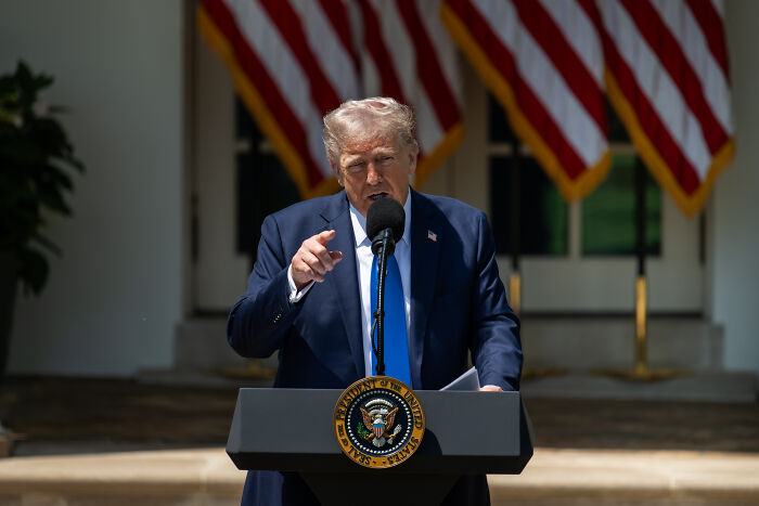 Donald Trump speaking at a podium with presidential seal, addressing first 100 days and mistakes amid flags.