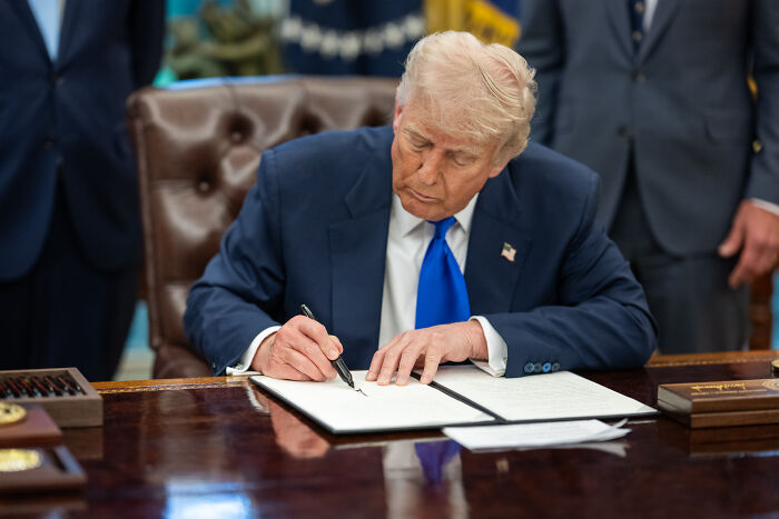 Donald Trump signing a document in the Oval Office related to policy on trans troops amid legal dispute.